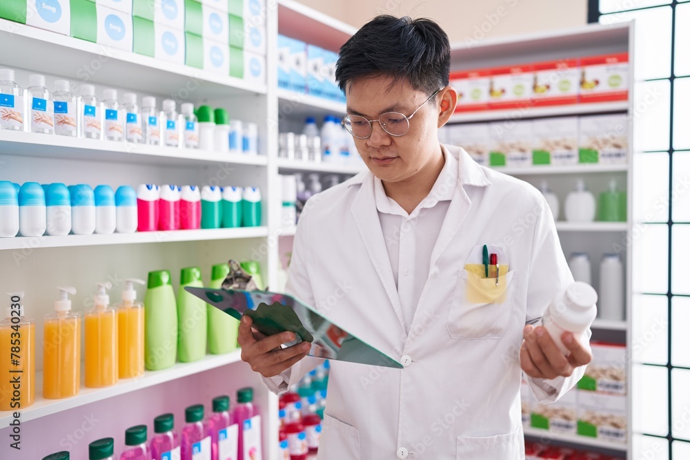 Young chinese man pharmacist holding pills bottle reading prescription at pharmacy