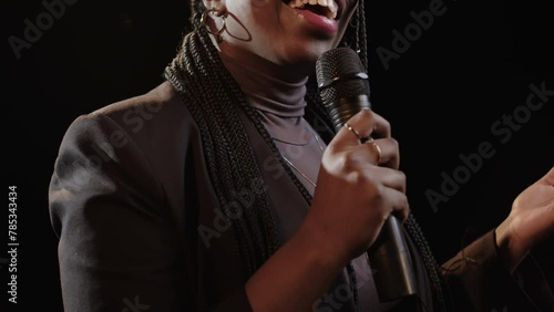 Chest up of young Black woman with braided hairstyle standing with microphone on dark stage and speaking while performing standup comedy monologue
