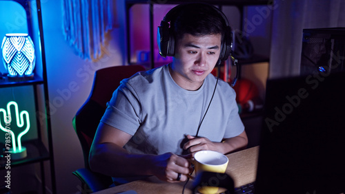 A young asian man with headphones in a gaming room lit by neon lights at night.