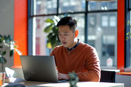 Focused Asian man working on laptop in a bright office.