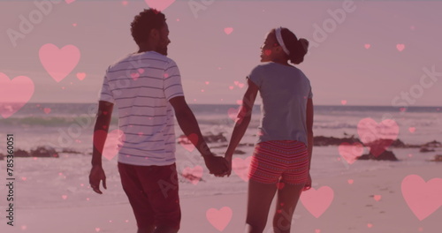 Multiple pink hearts floating against african american couple holding hands and walking on the beach