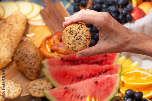 Close-up view of a hand holding a cookies with seeds against the background of laid out picnic food, selective focus. The concept of summer outdoor recreation on the weekend