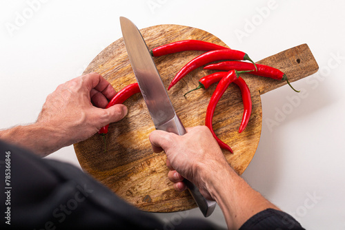 Top view of the hands of a male cook who cuts red chili peppers with a knife on a cutting board, flat lay. Episode of the cooking process