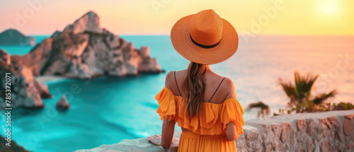 A woman in a yellow dress and a straw hat stands on the sea and rocks