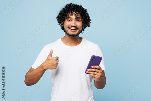 Young happy fun Indian man he wear white t-shirt casual clothes hold in hand use mobile cell phone show thumb up isolated on plain pastel light blue cyan background studio portrait. Lifestyle concept.