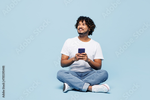 Full body young happy Indian man he wears white t-shirt casual clothes sits hold in hand use mobile cell phone isolated on plain pastel light blue cyan background studio portrait. Lifestyle concept.
