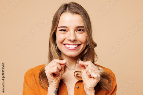 Young fun woman wear orange shirt casual clothes hold in hand invisible transparent aligners, invisalign bracer smile isolated on plain pastel light beige background studio portrait Lifestyle concept