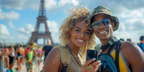 A woman is smiling and holding a cell phone in front of the Eiffel Tower. The scene is lively and joyful, with people around her enjoying the moment © olgakudryashova