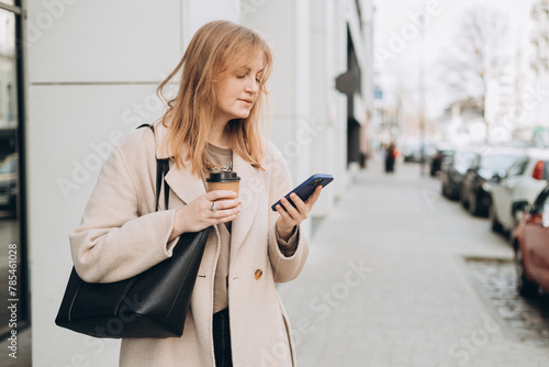 Beautiful smiling female with coffee cup using smartphone and standing outdoors. Phone Communication. Happy cheerful young woman walking on city street, Urban lifestyle concept. Traveler