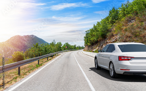 A white car drives along the highway against the backdrop of rocky mountains on a sunny day.
