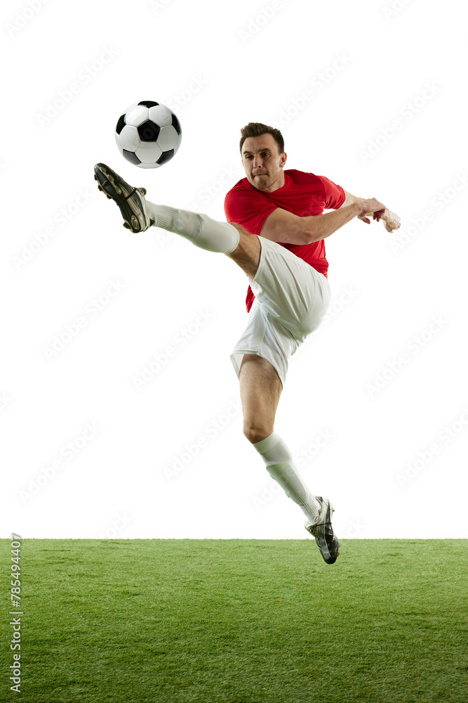 Full-length of young man in red and white uniform, football player hitting ball in a jump, training isolated on white background. Concept of professional sport, game, competition, tournament, action