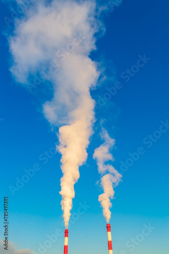 Two tall red and white smoke stacks are spewing smoke into the sky. The sky is clear and blue, with no clouds in sight
