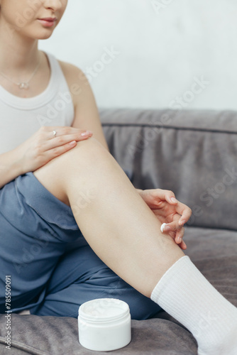 Portrait of young adult woman applying body lotion for skin on legs while sitting on couch at home