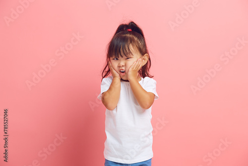 Puzzled little asian girl feels fear, holds her hands on cheeks, looks with great surprise and amazement at the camera standing on a pink isolated background