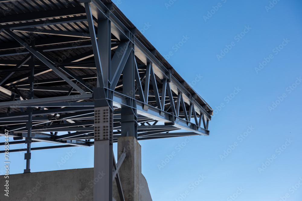 Fragment of a canopy. Metal beams on a concrete base support a metal roof against a blue sky - obrazy, fototapety, plakaty 