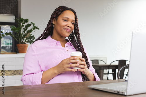 Biracial woman holding coffee, looking at laptop