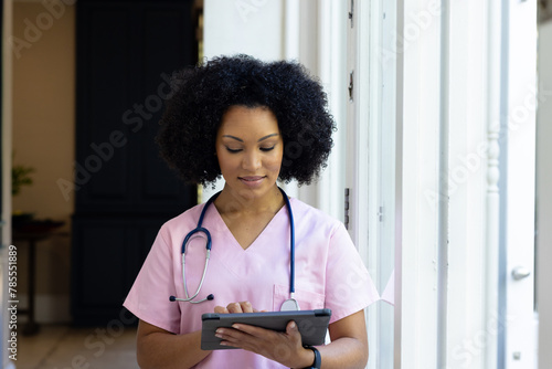 Young biracial female nurse wearing pink scrubs checking tablet at home