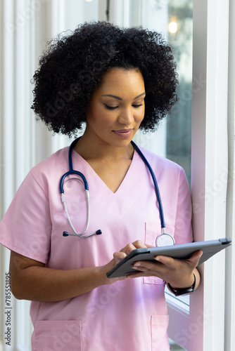 Biracial female nurse wearing pink scrubs reviewing notes on tablet at home