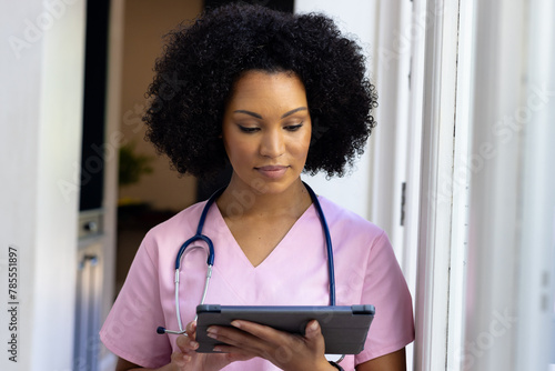 Young biracial nurse wearing pink scrubs checking tablet at home