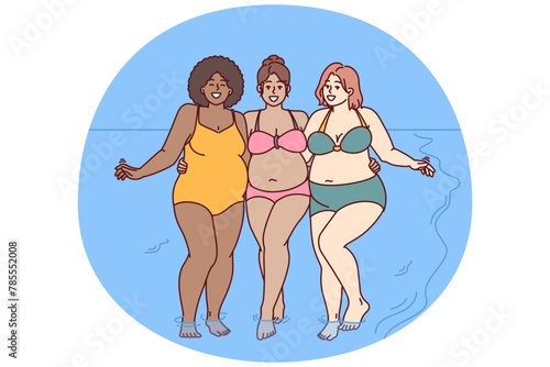 Cheerful plus size women dressed in swimsuit stand hugging on sunny beach near sea water and look at camera smiling. Diverse plus size girls for concept of no prejudice and body positivity