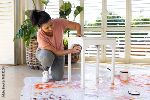 Mature biracial woman painting table at home in an upcycling project, wearing casual clothes