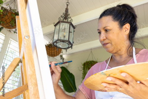 Mature biracial woman painting on canvas at home, focused on art