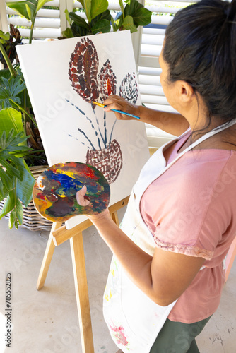 Mature biracial woman painting on canvas at home, wearing apron
