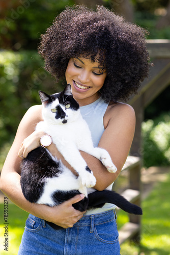 Biracial young woman holding black and white cat, both outdoors at home in garden