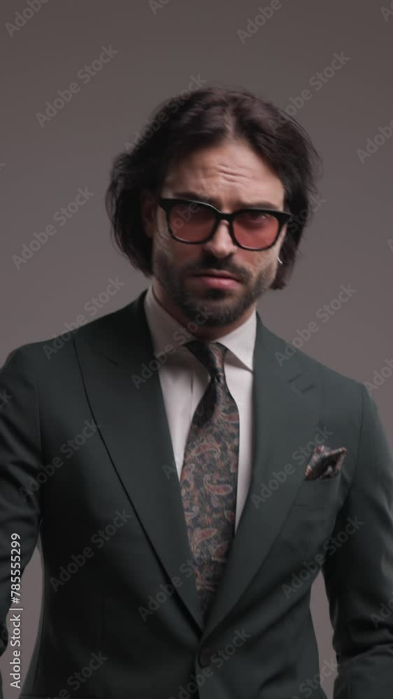 cool elegant man with sunglasses in suit looking to side and walking, confidently posing, adjusting suit and holding hands in pockets on grey background cool elegant man with sunglasses in suit looking to side and walking, confidently posing, adjusting suit and holding hands in pockets on grey background