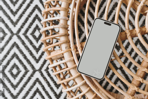 Mobile phone with blank screen on ornamental bamboo table and carpet. Flat lay, top view mock up with copy space. Bohemian, coastal, organic modern and tropical style