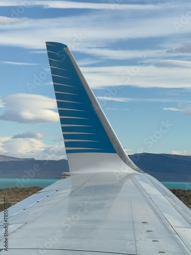 The tail of an airplane is seen in the distance, with a blue and white stripe. The sky is clear and blue, and the mountains in the background are visible. Concept of adventure and travel