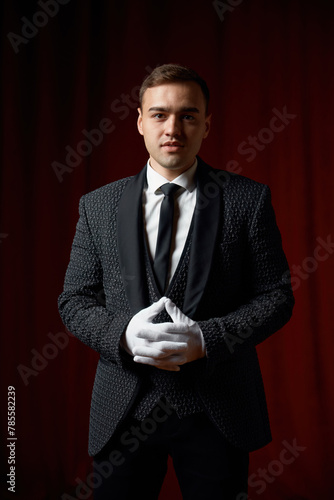 Magician wearing suit standing with cane on stage decorated red velvet curtain