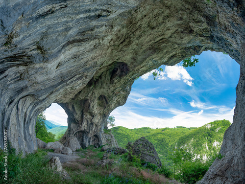Viewpoint above mountains through a cave, eroded in a calcareous cliff on a mountain side. The tunnel has its stone walls covered with moss. The arches in the cave are untied by a pillar. Carpathia