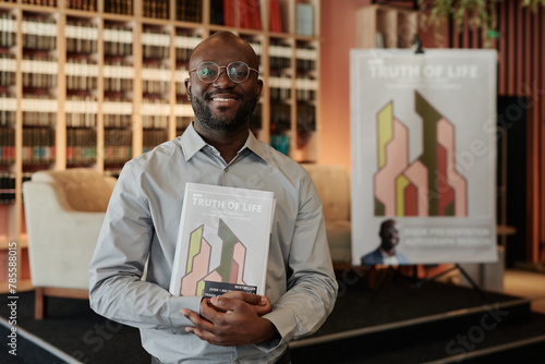 Young successful African American writer with his new book in hands looking at camera during presentation event in library