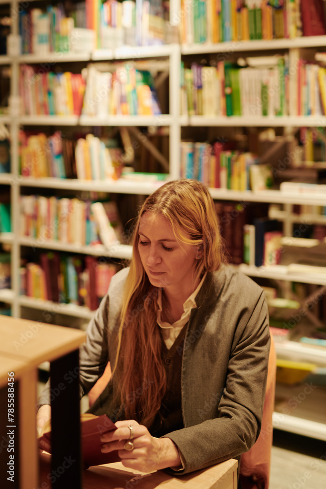 Young female bibliophile in smart casualwear reading book in library while sitting by desk against stack of bookshelves Young female bibliophile in smart casualwear reading book in library while sitting by desk against stack of bookshelves