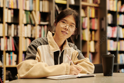 Successful Asian student in eyeglasses looking at camera while making notes in copybook during preparation for session photo