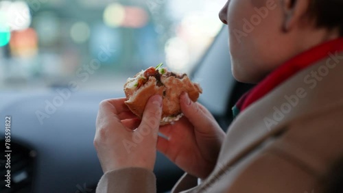 Lady consumes unhealthy fast food in car driving along town in cloudy weather. Lady sitting in car and relishing fast food. Lady knowingly indulges in fast food detrimental to health in car photo