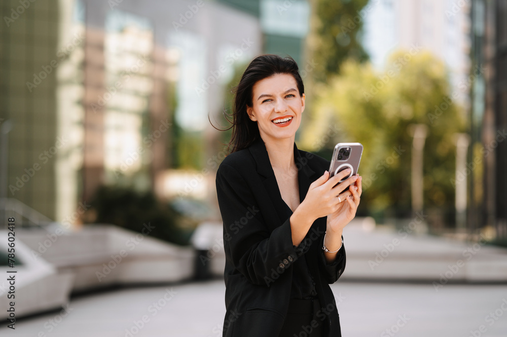 Young wide smiling woman wearing black suit is using her phone outdoors. Young wide smiling woman wearing black suit is using her phone outdoors.