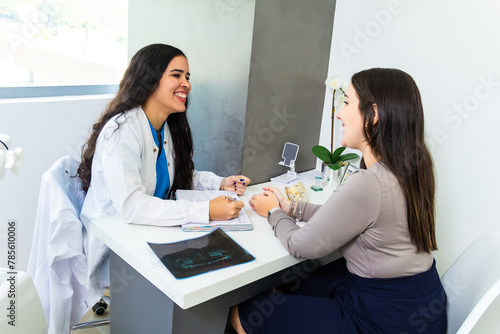 Young woman patient and latin dentist laughing together or smiling while sitting in office desk in a dentistry clinic