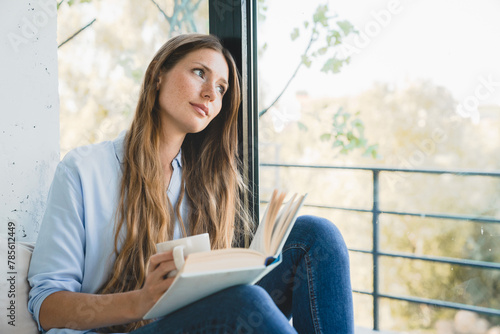 Dreamy young Caucasian woman reading a book, inspired by the plot sitting by the window at home. Pretty smart girl studying learning, creating new ideas indoors