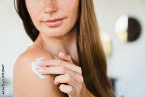 Cropped shot of young pretty woman applying body cream serum on shoulder indoors. Spa body hair treatment, beautification concept. Moisturizing effect photo