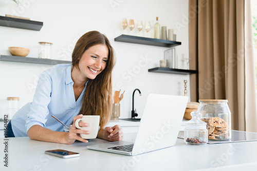 Smiley young freelancer working remotely on laptop while having breakfast at home kitchen. Office worker employee doing project, watching webinar, e-learning, checking emails