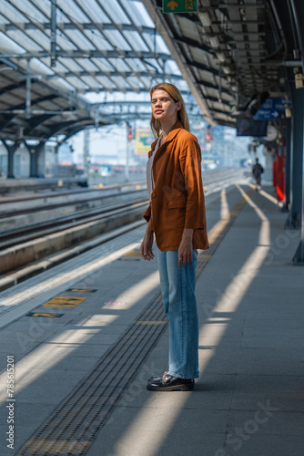 A contemplative young woman stands alone on a sunny train station platform, seemingly waiting for the next train.