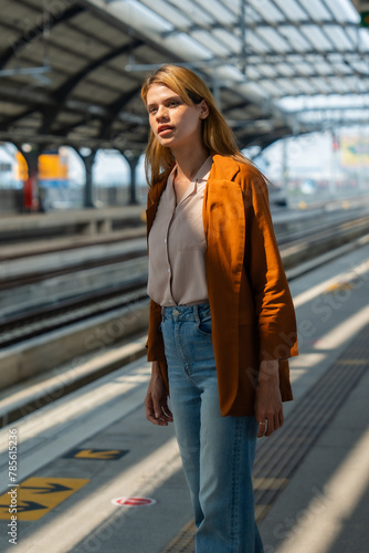 A contemplative young woman stands alone on a sunny train station platform, seemingly waiting for the next train.