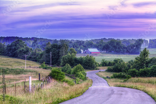 Valley Scene.  A winding country road meanders through a tranquil rural landscape, leading to a red-roofed barn in the distance. The scene is enveloped in the soft hues of dusk, with a blend of purple