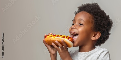 Happy Afro-American Boy With Mouth Open Eating a Hot Dog With Mustard  On a Gray Background With Copy Space  Studio Photo