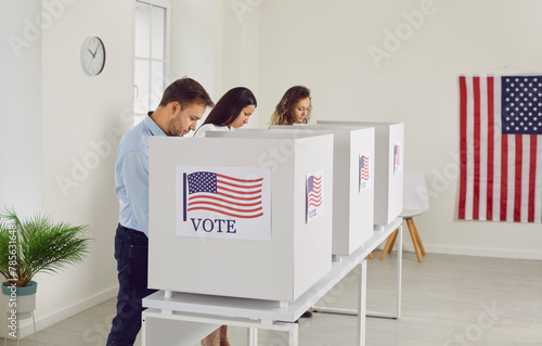 Concentrated people are reading information on election ballots before casting their votes. Young men and women vote anonymously in voting booths at polling station decorated with American flags. photo