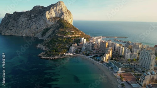 Beautiful sunset over Calpe city and Peñón de Ifach mountain. Drone backwards and panning right. Sunset colours reflecting on buildings and mountain. Travel destination in Spain, Alicante Province.