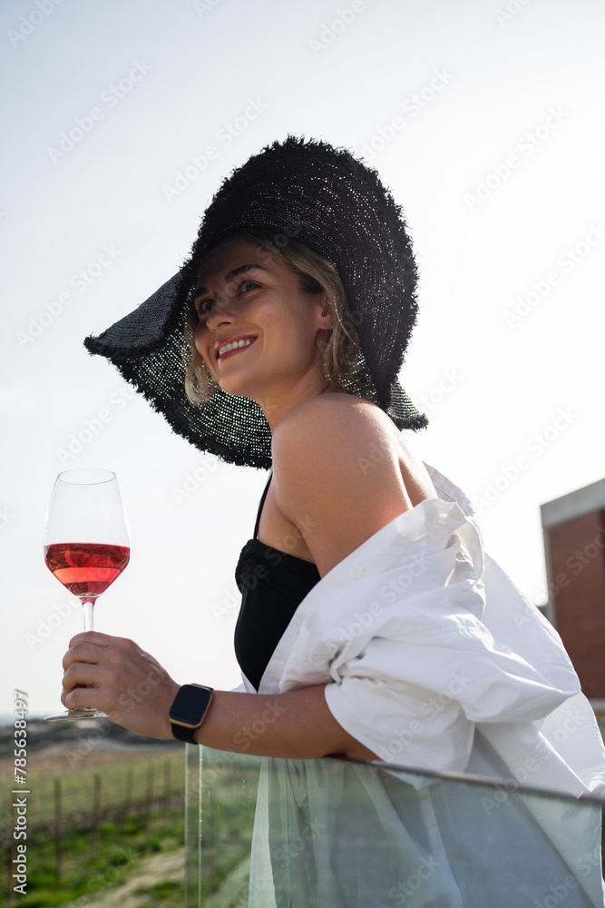 Fototapeta premium Woman in elegant attire and a wide-brimmed hat enjoys a glass of rose wine at a vineyard
