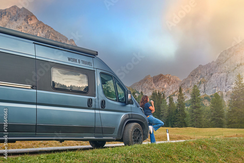 Attractive middle-aged woman leans against motorhome, sipping coffee with Dolomites peaks behind. 
